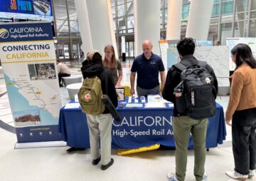 Two employees stand behind a table with a cloth reading California High-Speed Rail Authority, presenting on the project to three members of the public.
