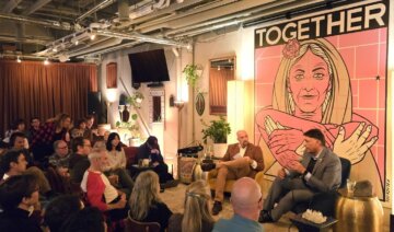A seated audience fills a small event space, facing towards two men seated at the front of the room holding a discussion. The room includes warm lighting, plants, side tables, and a colorful mural that reads “Together” behind the two men.