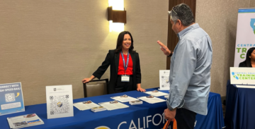 A woman in professional clothing behind a California High-Speed Rail Authority informational table speaking to a man. There are factsheets and other resource sheets on the table. There is a wall light behind her.