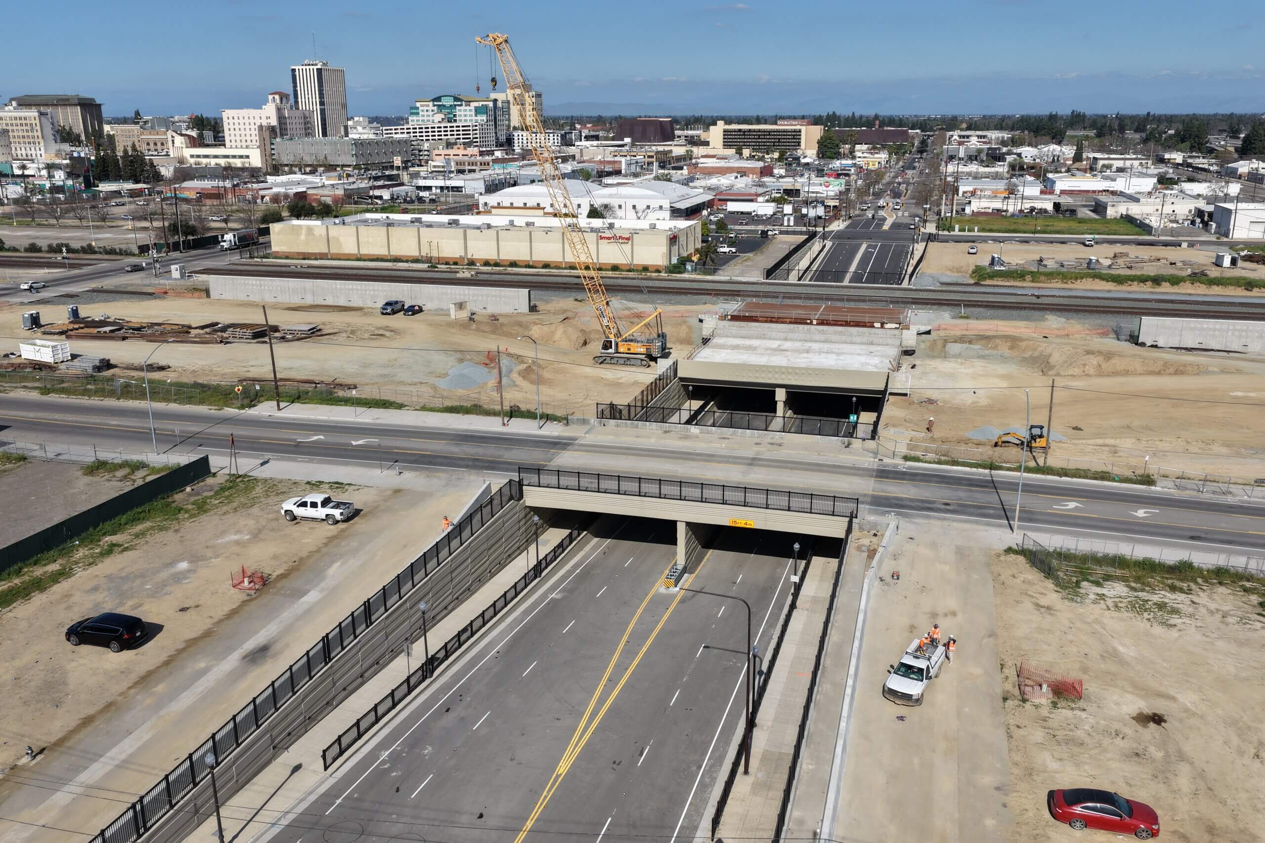 A drone shot, high and at an angle, of the Cesar Chavez Boulevard underpass grade separation project, looking approximately from west to east, the skyline of downtown Fresno in the background. A cloudless sky above the horizon; urban concrete, wires, and bare dirt below it.