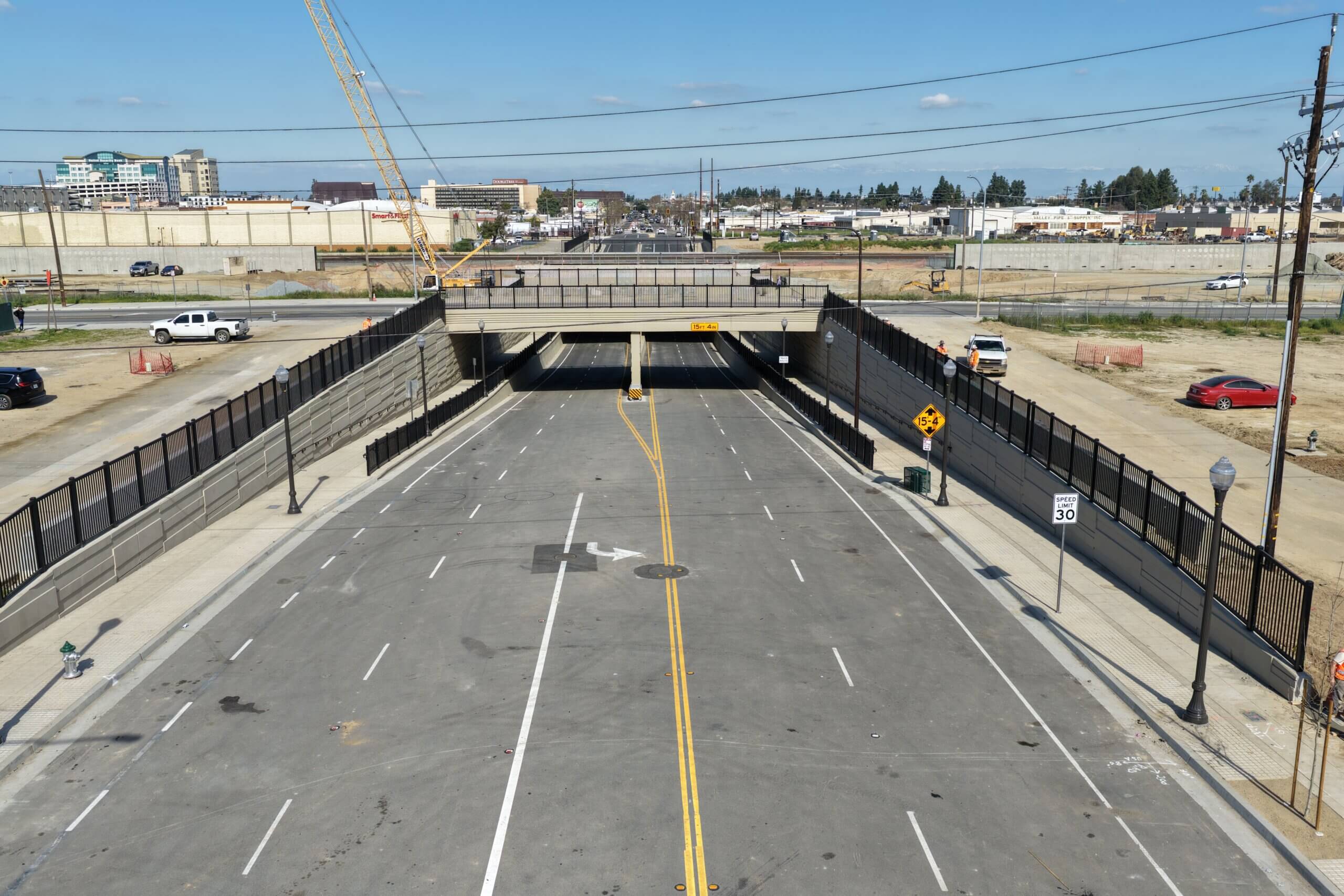 A drone shot, low, of the Cesar Chavez Boulevard underpass grade separation project, looking from west to east. A cloudless sky above the horizon; urban concrete, wires, and bare dirt below it.